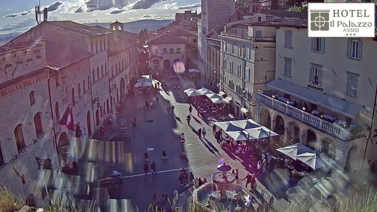 A sun-drenched town square with a fountain at its center is surrounded by stone buildings, with people strolling and dining under outdoor umbrellas.