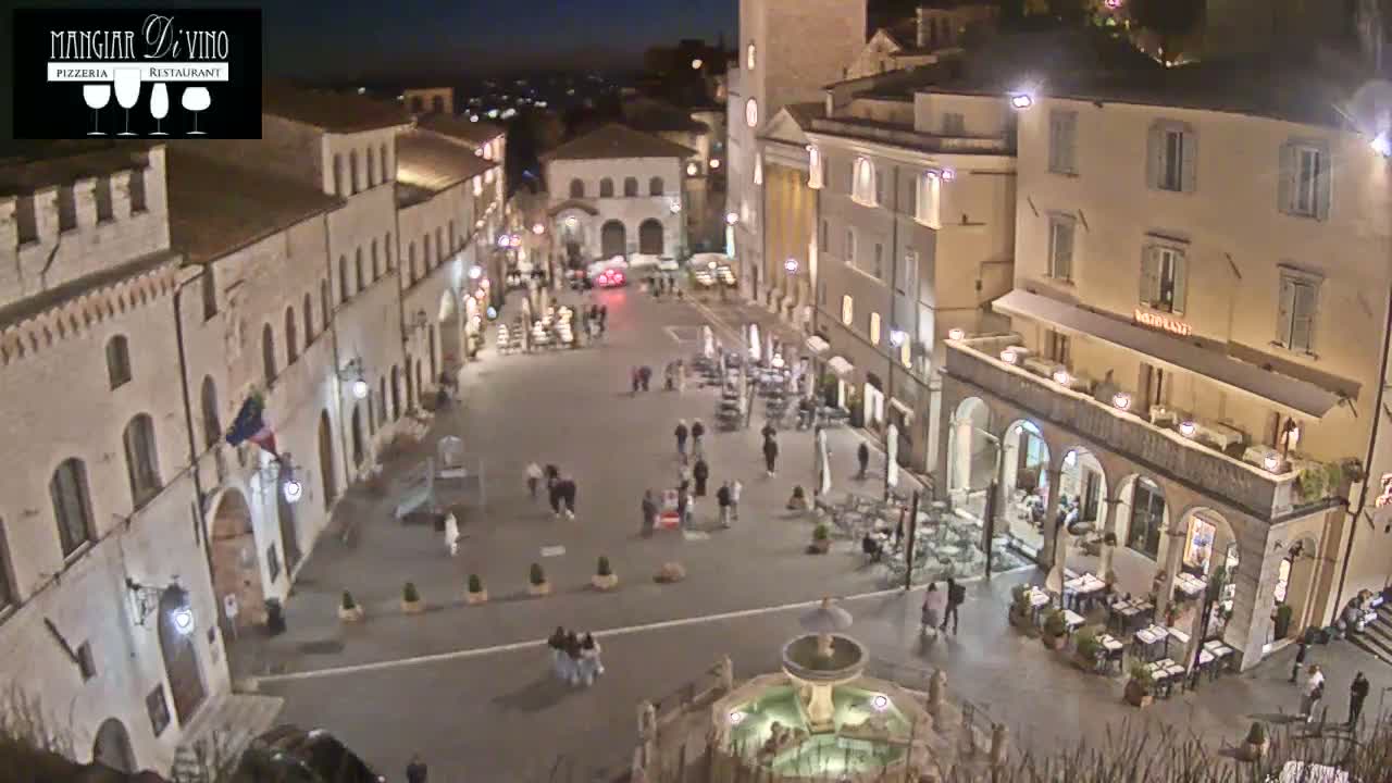 A nighttime aerial view of a town square with several buildings, a fountain, and people walking around.