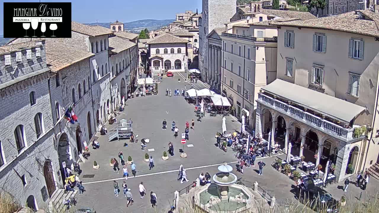 A sunny day in a large town square in Italy, surrounded by historic stone buildings, with people sitting at outdoor cafes and walking around a central fountain.
