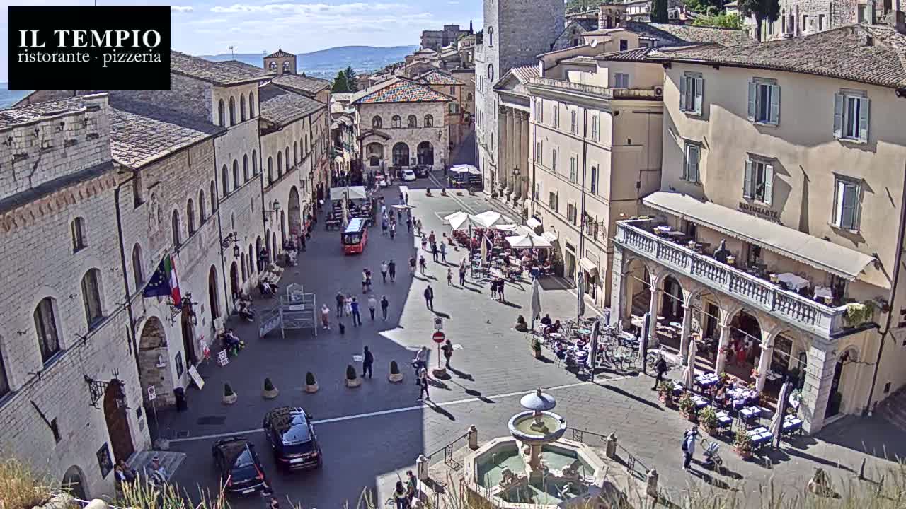 A sunny town square, filled with people at outdoor cafes surrounding a central fountain, is nestled amongst historic stone buildings.
