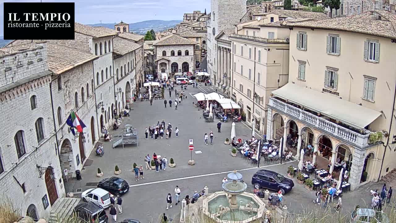 A town square, under an overcast sky, is filled with people and cars, centered around a fountain.