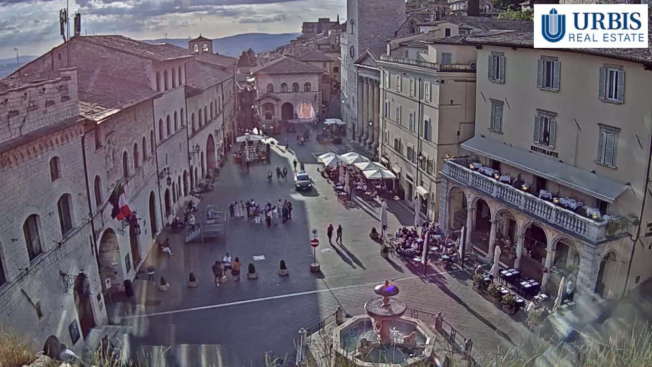 A sunlit town square with several buildings, outdoor seating areas, a fountain, and a scattering of people.