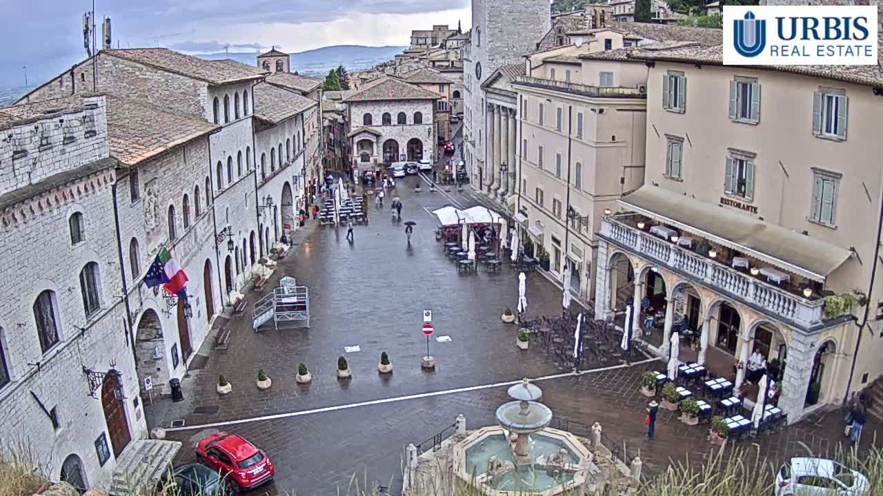 A wet, stone town square in Italy with several buildings, a fountain, outdoor seating, and a few pedestrians under an overcast sky.