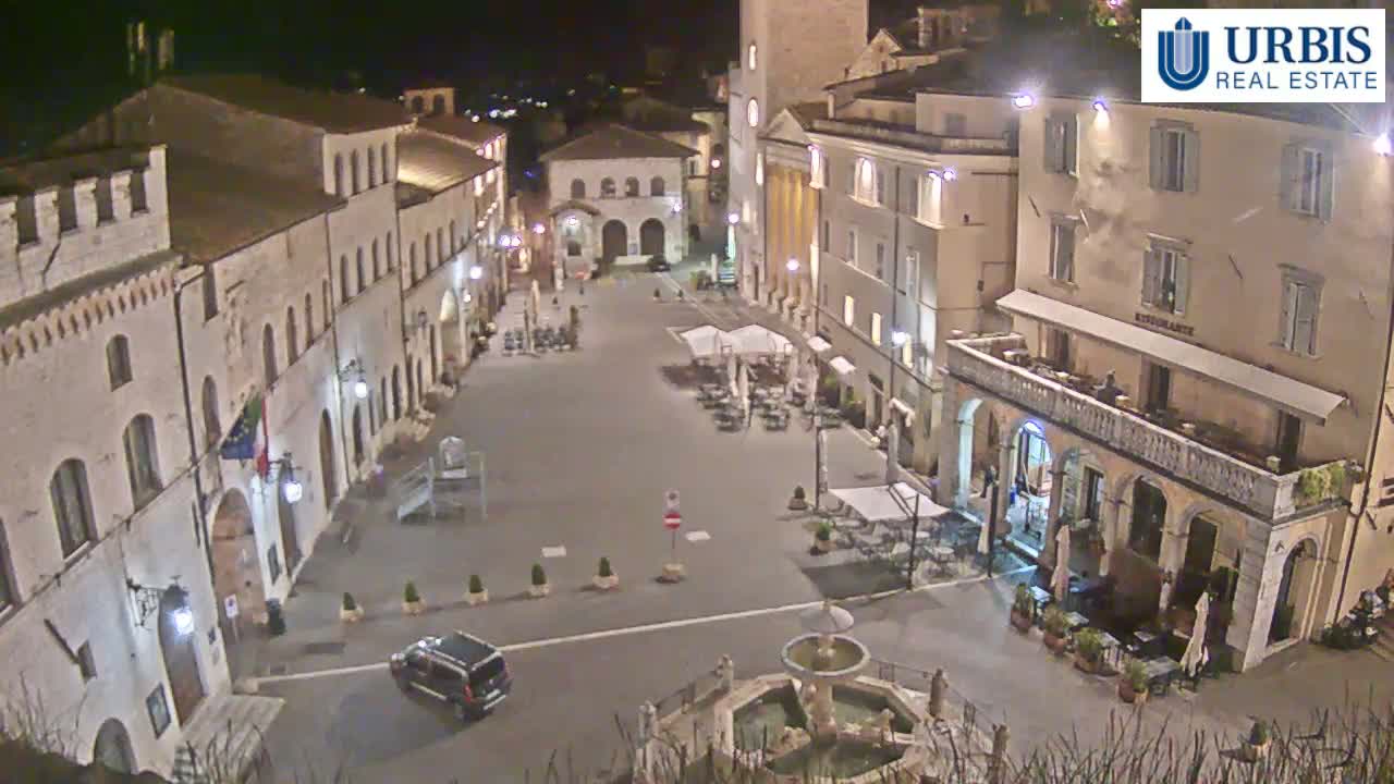 A mostly empty town square at night, surrounded by illuminated buildings, features a fountain in the center.
