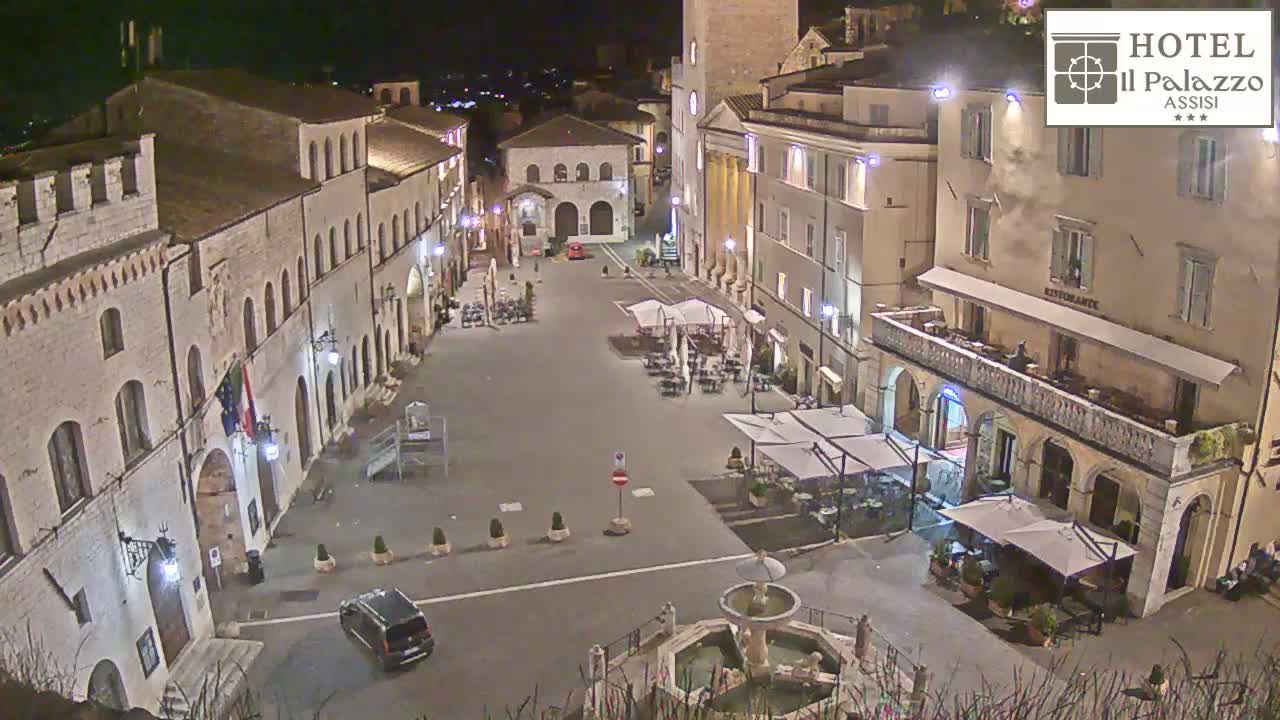 A nighttime aerial view of a town square in Italy, featuring several buildings, outdoor seating areas, a fountain, and a vehicle parked near the edge of the square.