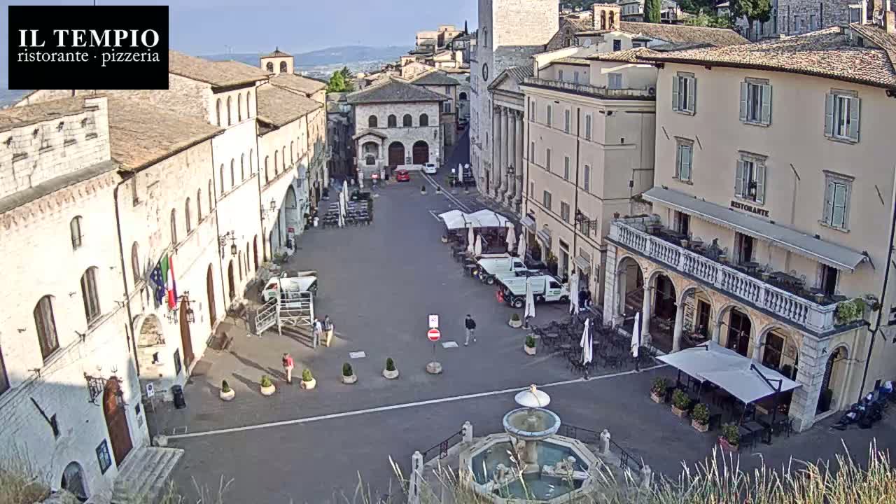 A sunny view of a town square in a European city, with several buildings surrounding the square, a fountain in the center, and some people and parked vehicles.