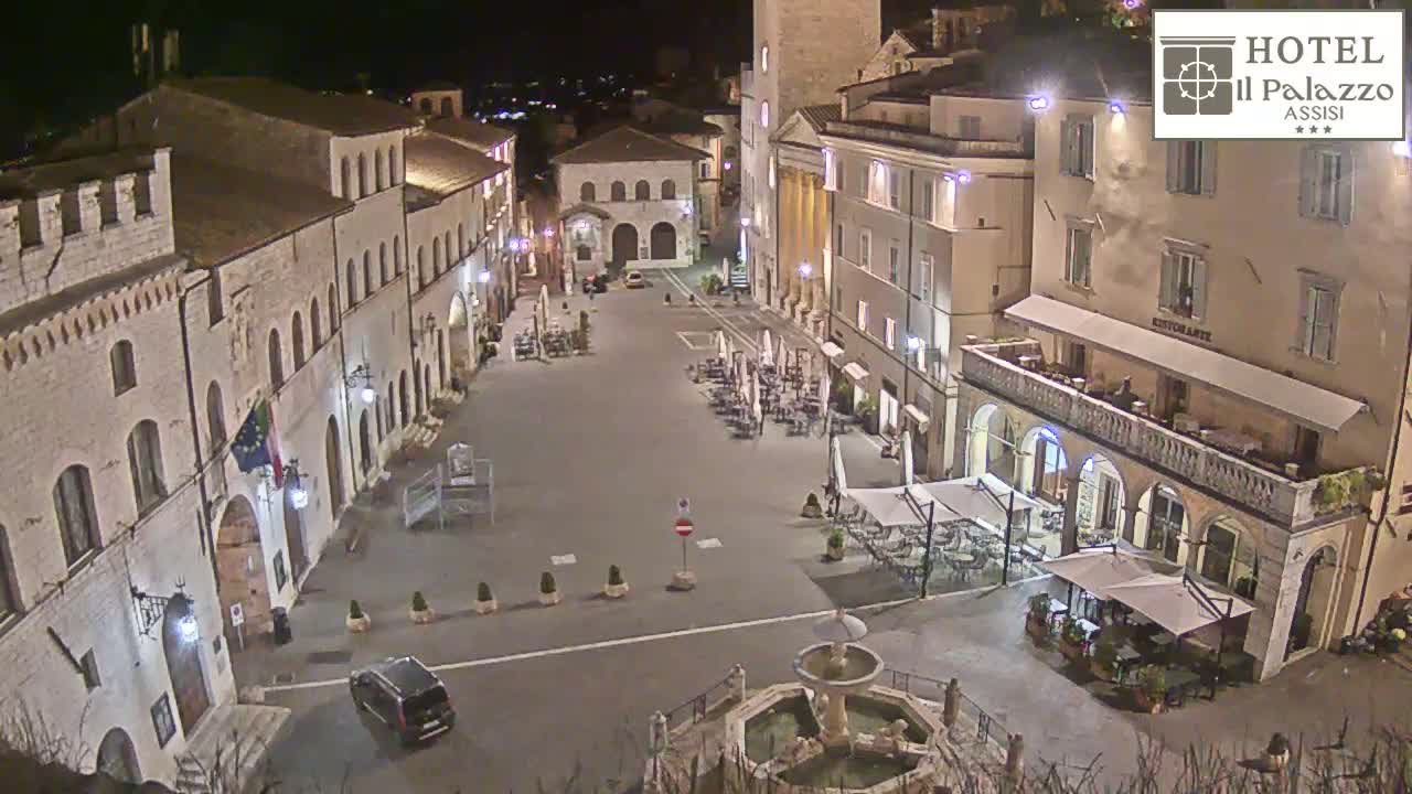 A nighttime view of a town square, with several buildings illuminated, outdoor seating areas at cafes, and a fountain in the center.