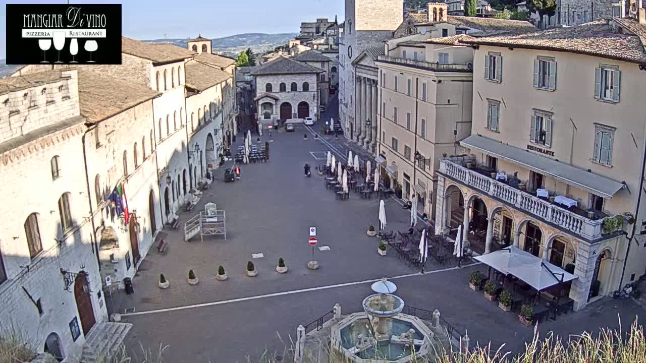 A mostly empty town square with a fountain at its center, surrounded by buildings under a clear, sunny sky.
