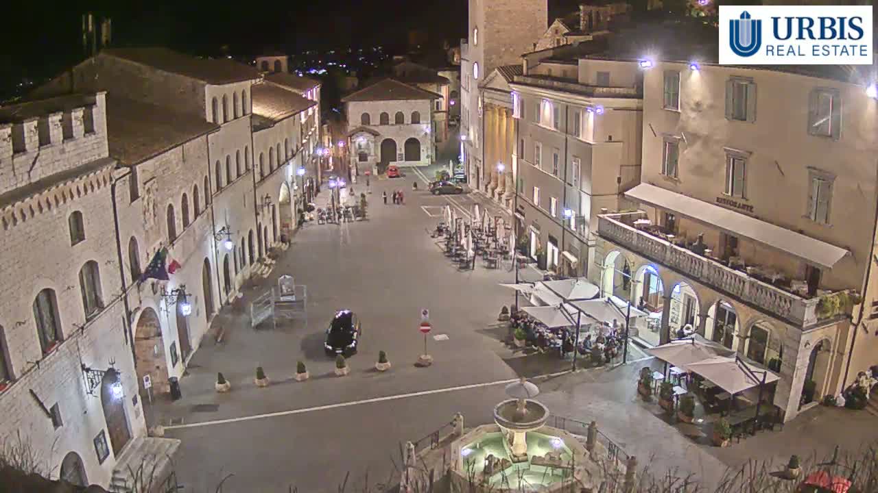 A nighttime view of a town square in a European-style town, with several buildings surrounding the square and outdoor seating at restaurants.