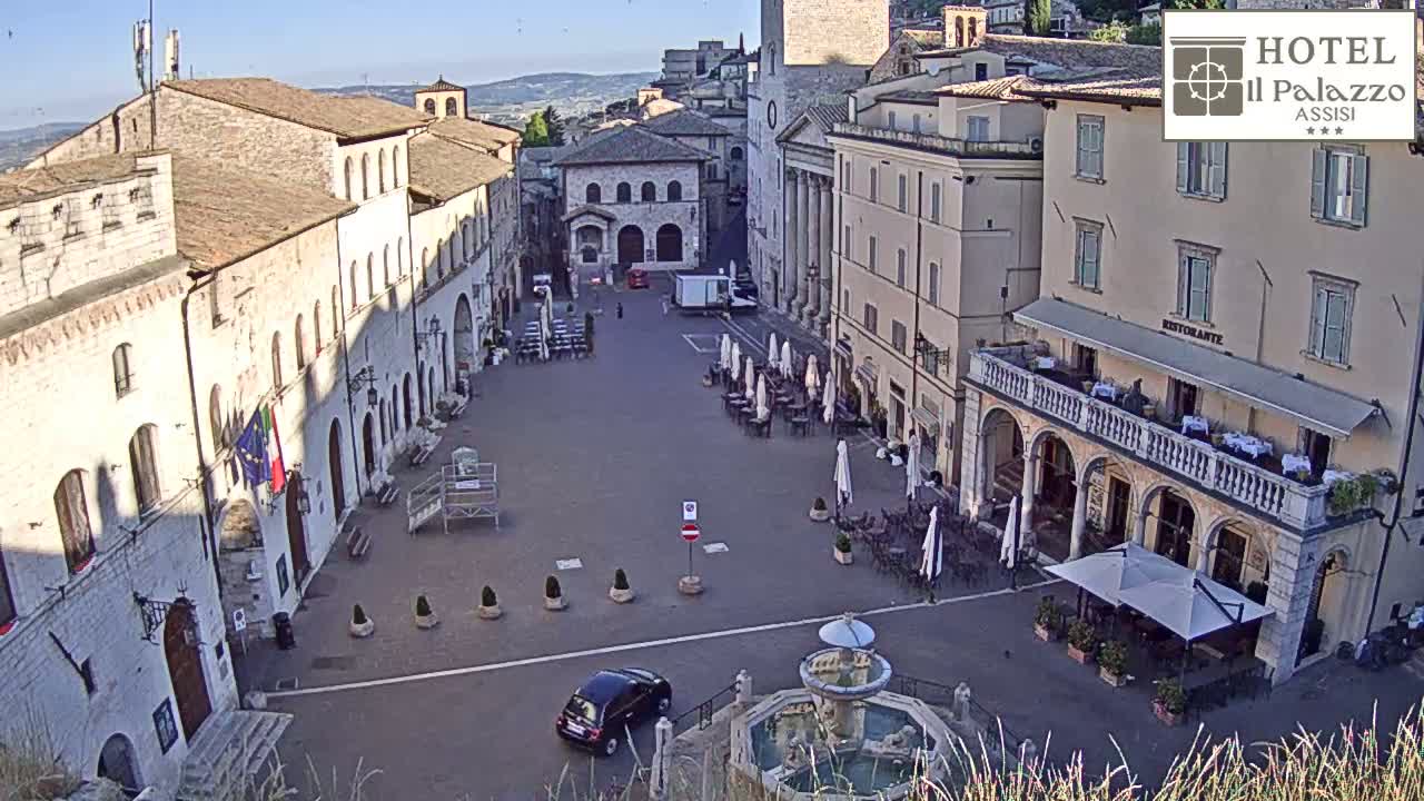A mostly empty town square in bright sunlight, featuring several buildings, outdoor seating areas, a fountain, and a single car.