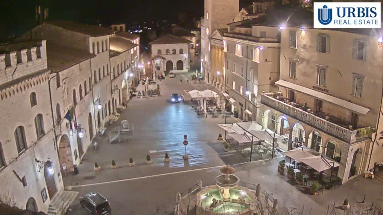 A nighttime view of a town square, featuring several buildings with outdoor seating, a fountain, and a few cars.