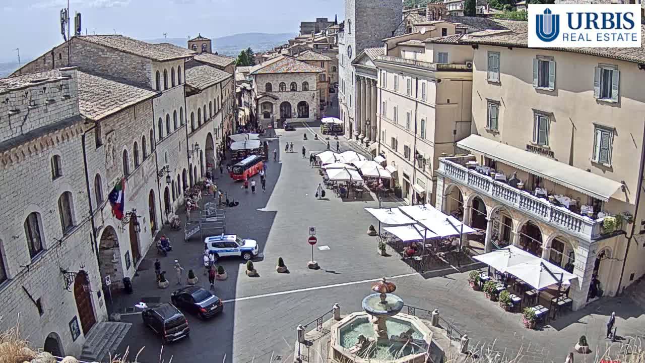 A sunny town square in Italy, filled with outdoor seating at cafes, a few cars, and some pedestrians, features a central fountain.