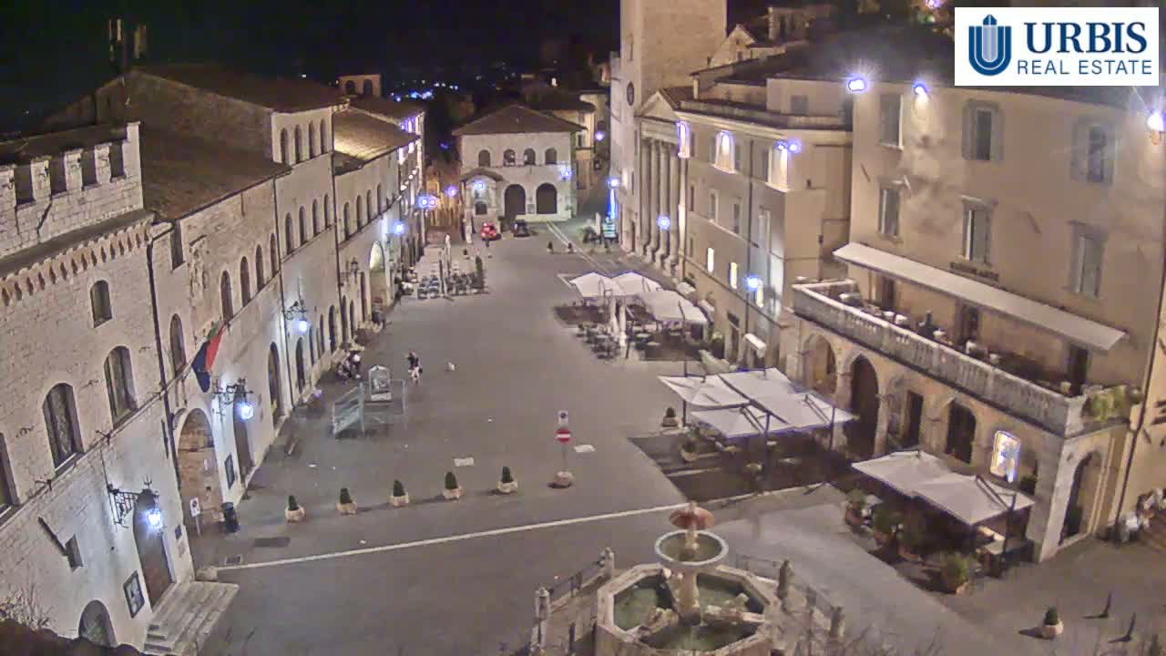 A nighttime view of a town square with several buildings, a fountain, and outdoor seating areas under umbrellas.
