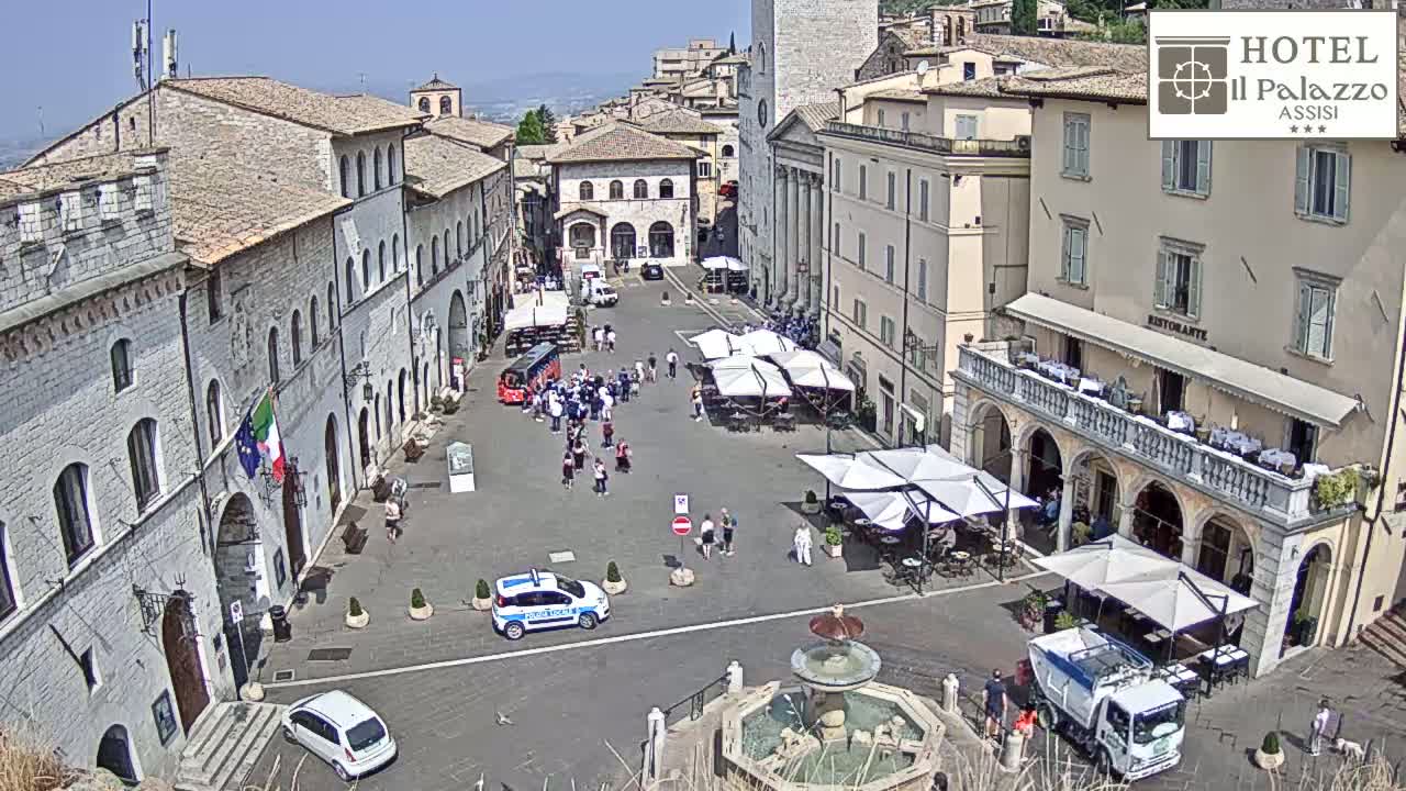 A sunny day reveals a town square in a historic Italian town, with several people, cars, and outdoor seating areas around a central fountain.