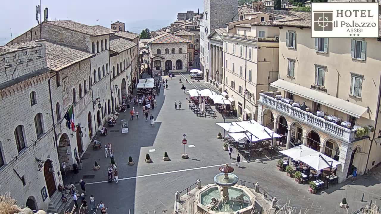 A sunny day reveals a large, open town square in a historic European city, featuring stone buildings, outdoor cafes, a fountain, and people walking about.