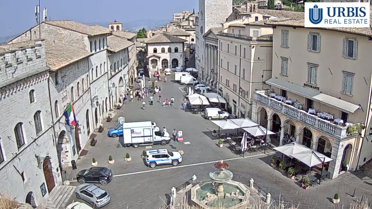 A sunny town square in Italy, surrounded by historic buildings, features a fountain, parked cars, and outdoor seating at a restaurant.