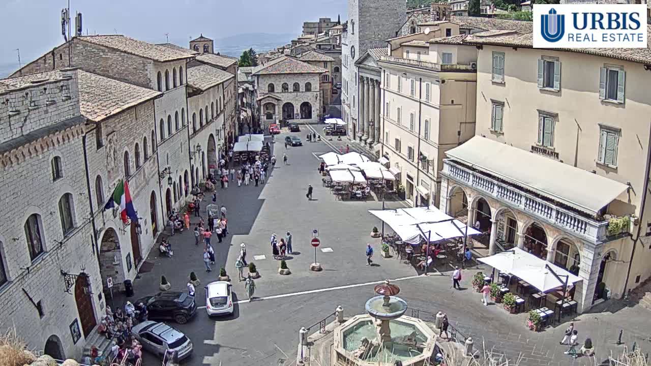 A sunny day reveals a town square in a European town, surrounded by stone buildings with outdoor seating areas and a fountain in the center.