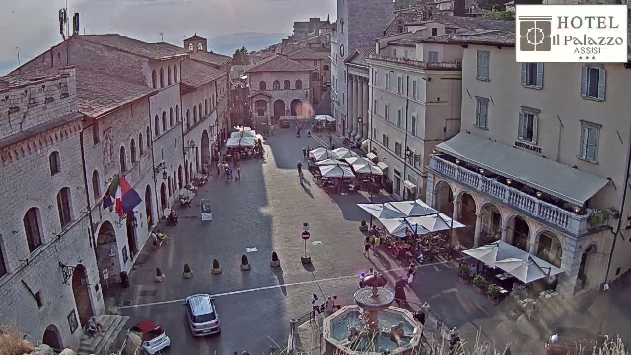 A sunlit town square in Italy, featuring several buildings, outdoor cafes with umbrellas, a fountain, and cars parked along the edges.