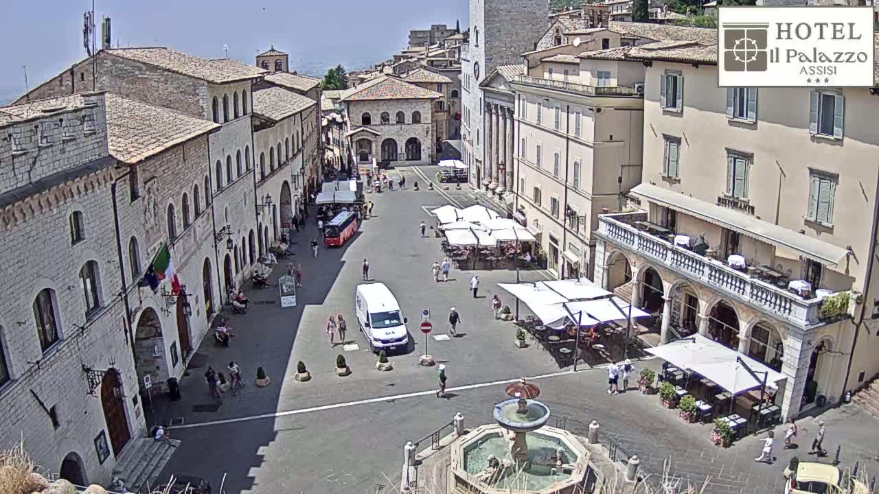 A sunny town square in Italy, filled with outdoor cafes and a central fountain, is surrounded by historic stone buildings.