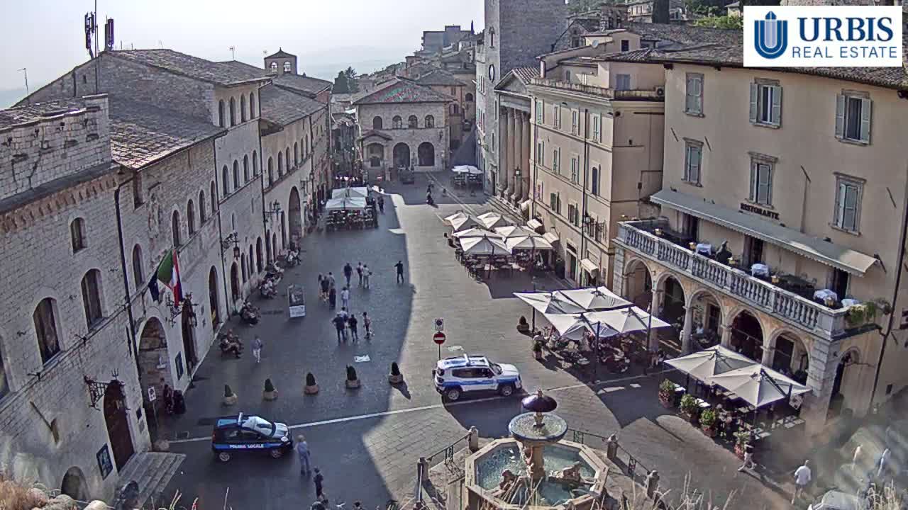 A sunny day in a town square with several buildings, outdoor cafes, a fountain, and a few parked cars.