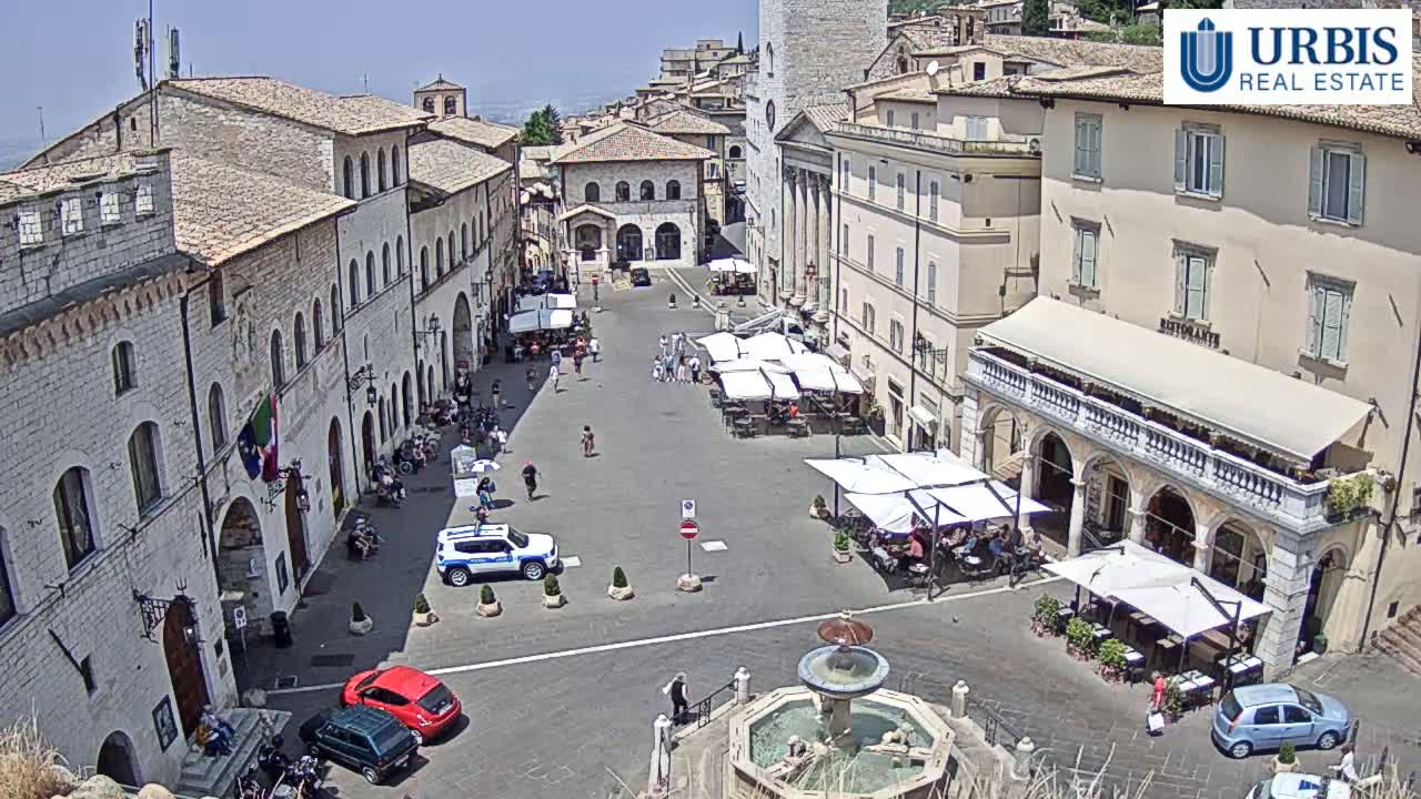 A sunny town square with stone buildings, outdoor seating at cafes, a central fountain, and a few cars parked.