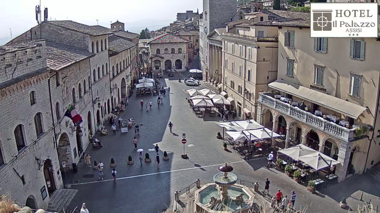 A sunny town square, filled with people, outdoor seating, and a central fountain, is surrounded by historic stone buildings.