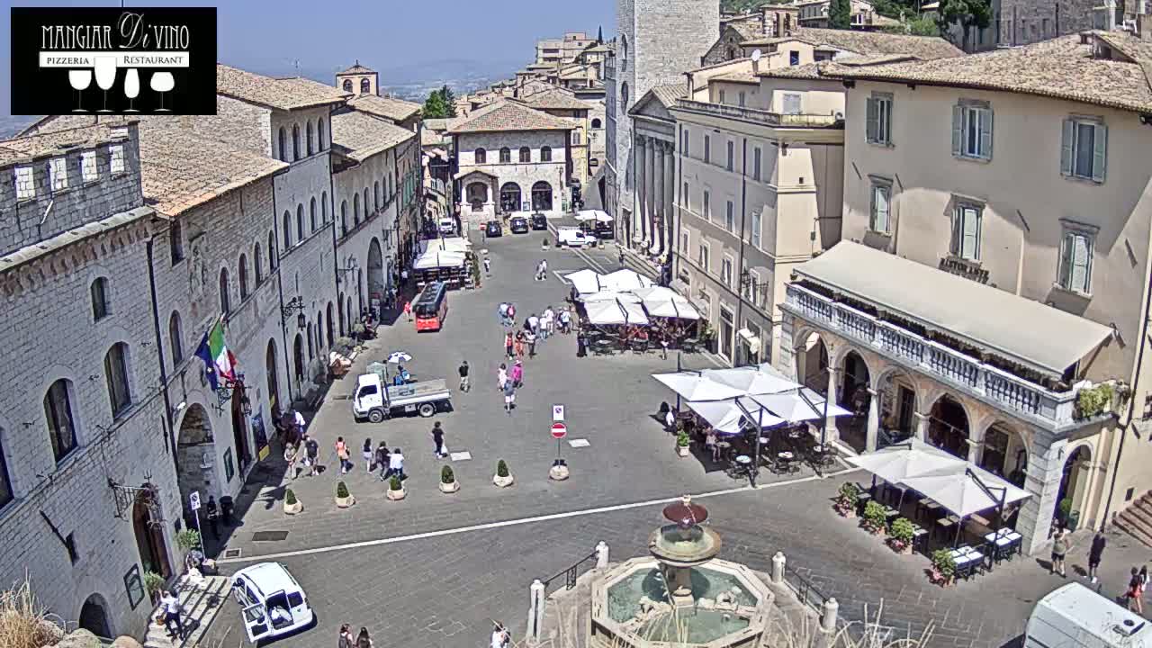 A sunny town square, surrounded by buildings, features outdoor seating and a central fountain with people walking around.