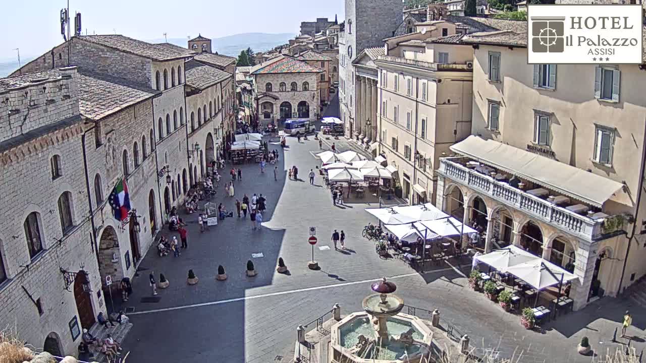 A sunny, high-angle view of a town square with several buildings, outdoor seating areas, a fountain, and people walking around.