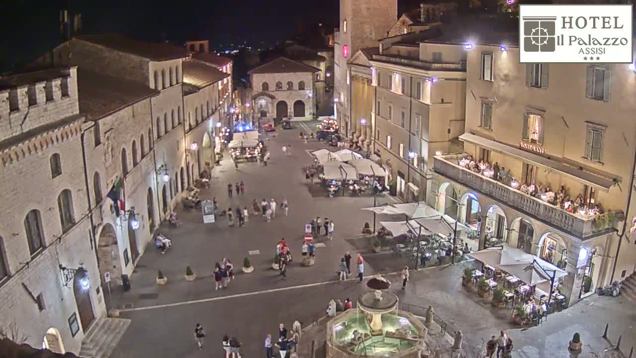 A sunny, high-angle view of a town square with several buildings, outdoor seating areas, a fountain, and people walking around.
