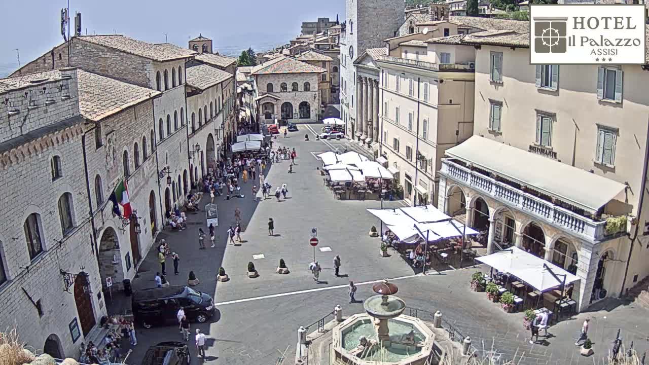 A sunny day in a European town square, with several people walking around and sitting at outdoor cafes, surrounded by old stone buildings and a central fountain.
