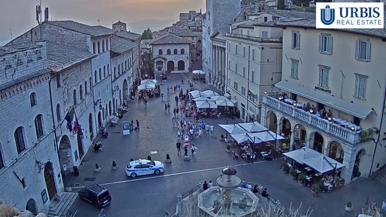 A town square filled with people sitting at outdoor cafes, under shaded umbrellas, near a fountain, during the evening.