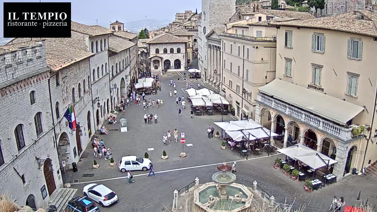 A high-angle view shows a town square in a sunny, possibly warm climate, with several outdoor cafes and a fountain in the center, surrounded by stone buildings.