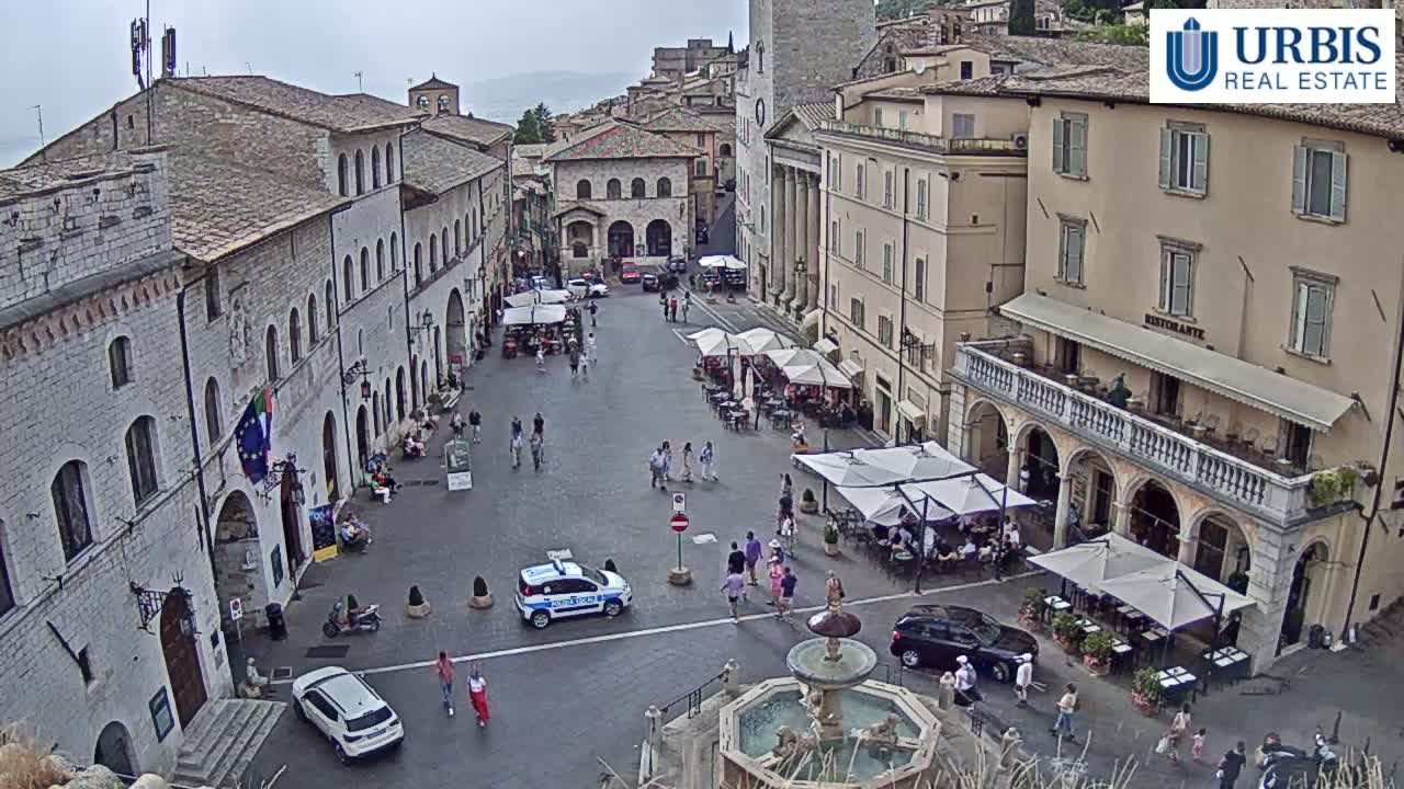 A mostly sunny town square in Italy is filled with people sitting at outdoor cafes and walking around, centered on a fountain, with older stone buildings surrounding it.