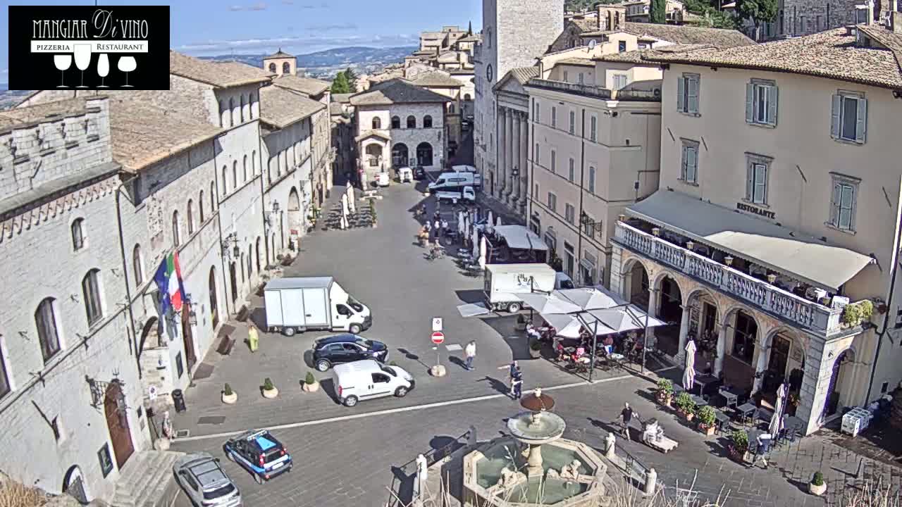 A sunny day in a town square, surrounded by old stone buildings, with cars parked, people walking, and outdoor seating at restaurants.