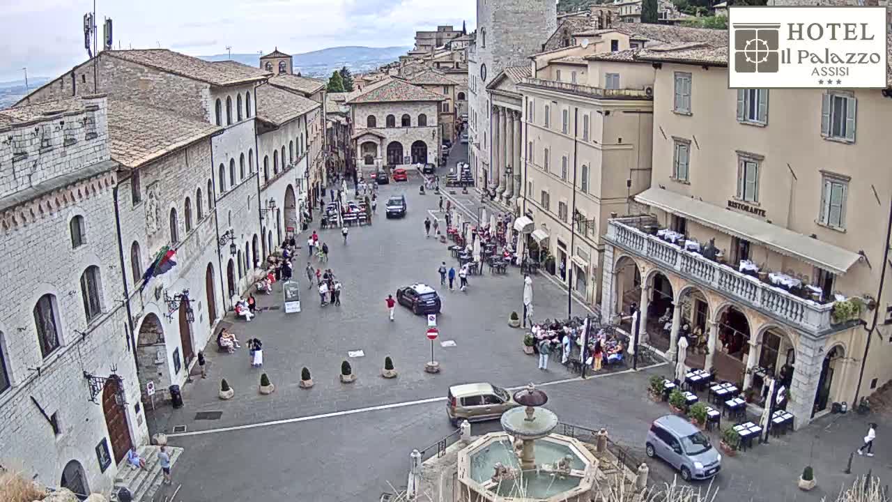 An overcast day reveals a town square in a European city, with people sitting at outdoor cafes surrounding a fountain, and cars parked near stone buildings.