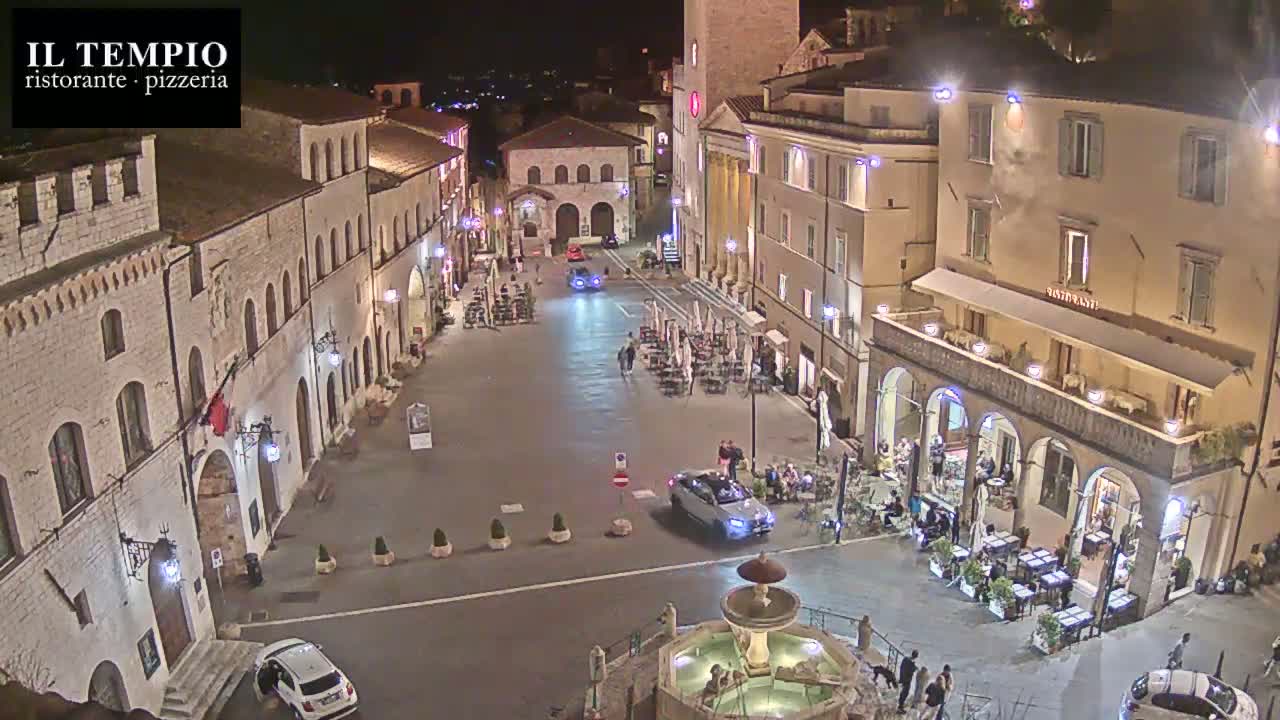 A nighttime view of a town square with several buildings, a fountain, cars, and people sitting at outdoor cafes.