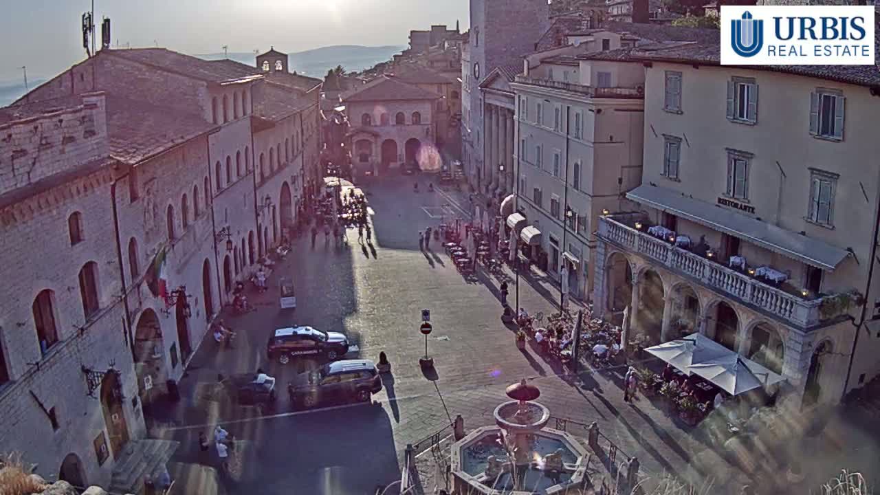 A sunlit town square with several buildings, a fountain, and people sitting at outdoor cafes, some cars parked nearby.