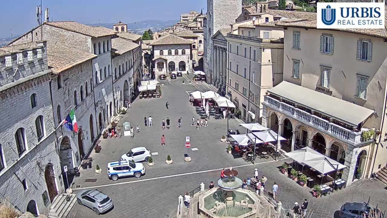 A sunny town square in Italy is filled with several outdoor cafes, a fountain, and a few pedestrians, surrounded by ancient stone buildings.