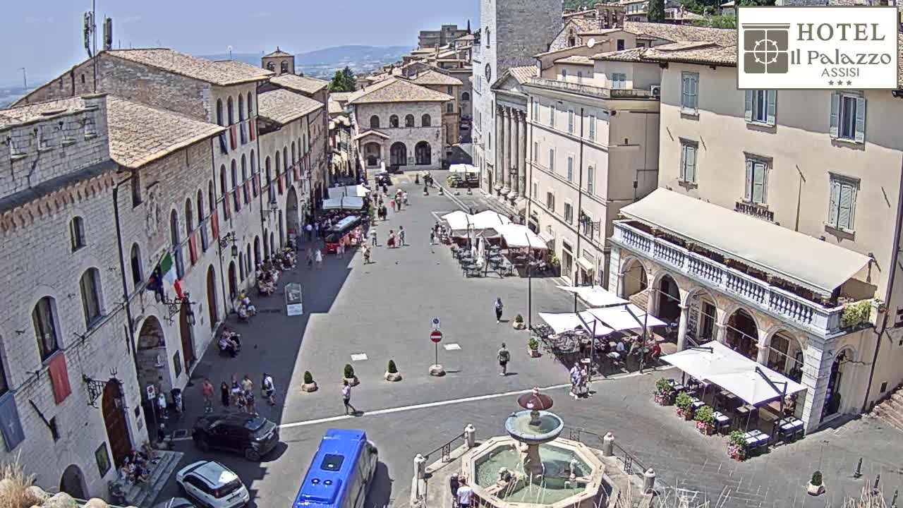 A sunny day in a town square filled with people, outdoor seating, and a fountain, surrounded by historic buildings.