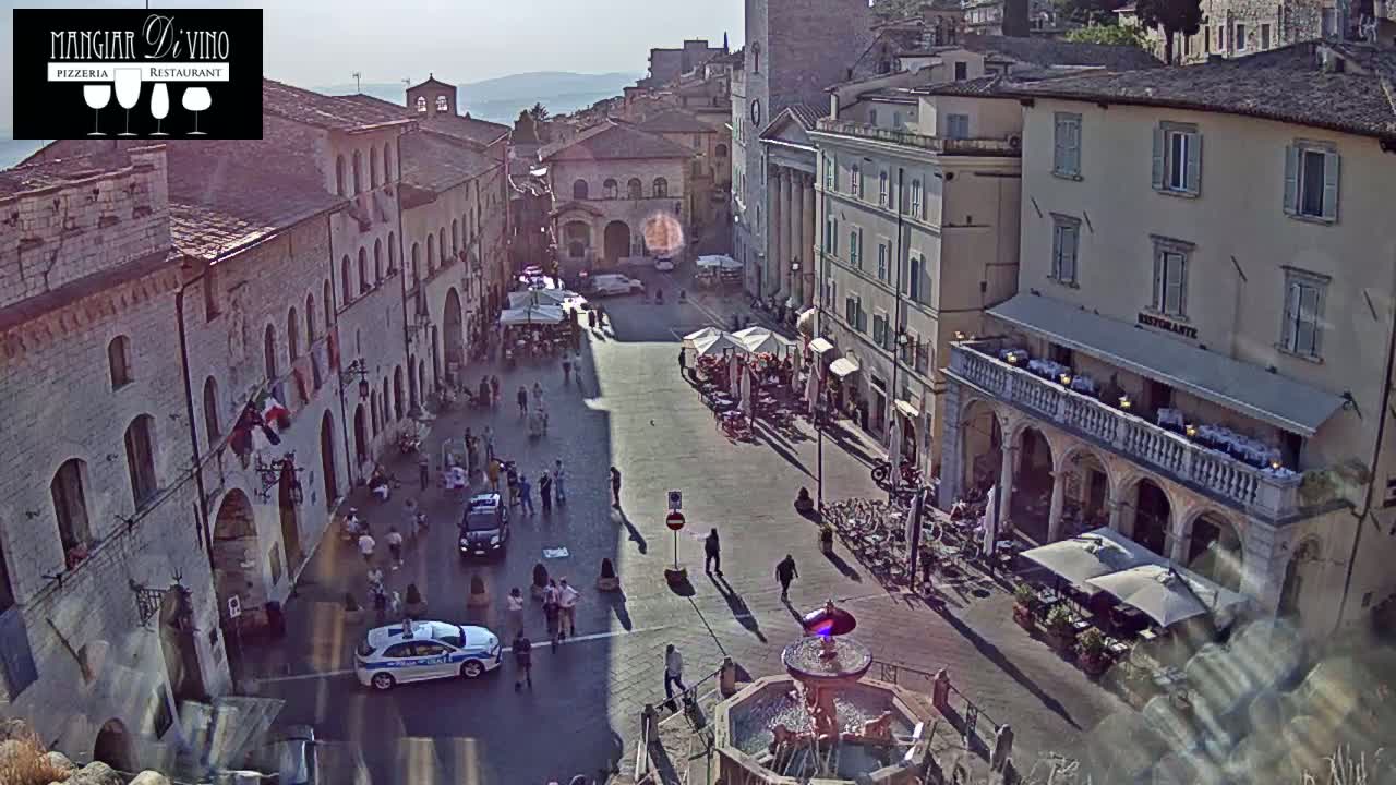 A sunny, high-angle view of a town square with several buildings, outdoor seating, pedestrians, and two police cars.