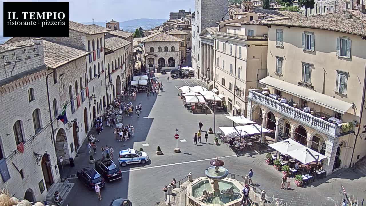 A sunny town square with a fountain in the center, surrounded by light-colored buildings with outdoor cafes and people walking around.