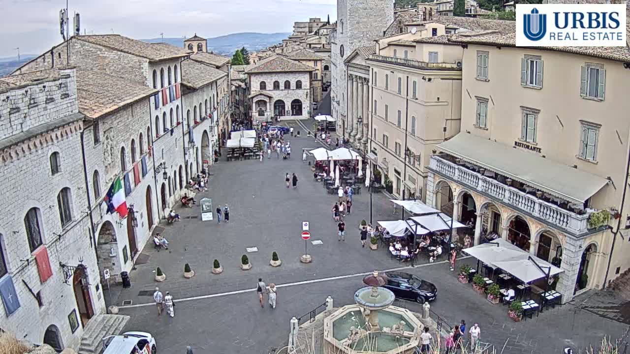 A town square in Italy, under a partly cloudy sky, is filled with people at outdoor cafes surrounding a fountain.