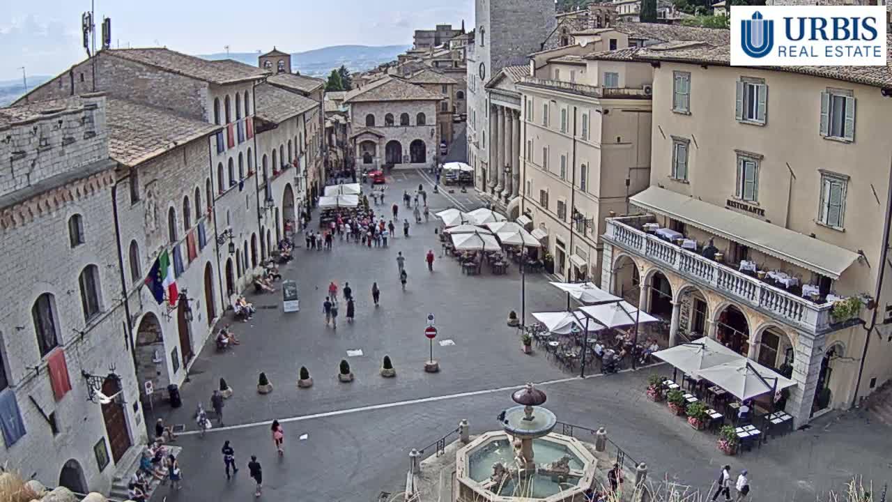 A large, paved town square with several buildings surrounding it, outdoor seating areas, and a fountain in the center, under a mostly clear sky.
