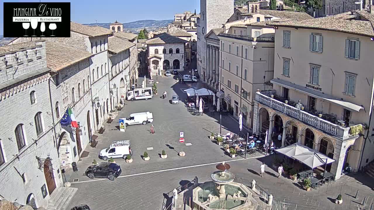 A sunny town square in Italy is surrounded by historical buildings, with a fountain at its center and several parked vehicles and outdoor seating areas.