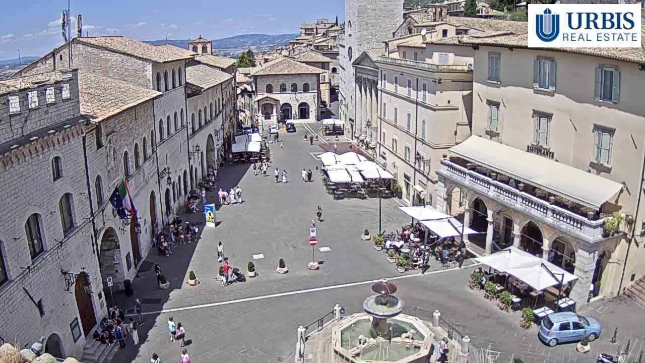 A sunny town square in Italy is surrounded by historical buildings, with a fountain at its center and several parked vehicles and outdoor seating areas.