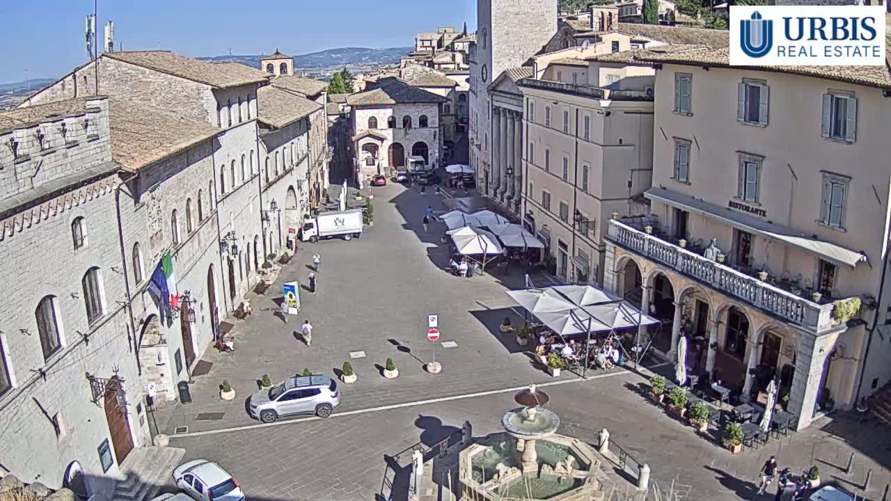 A sunny town square in Italy is surrounded by aged stone buildings, with outdoor seating at a restaurant and a fountain at the center.