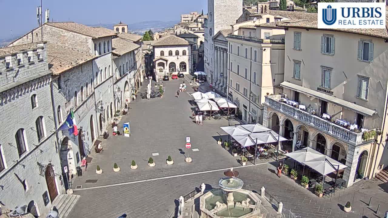 A sunny day reveals a large town square with several buildings, outdoor seating areas under umbrellas, and a fountain in the center.
