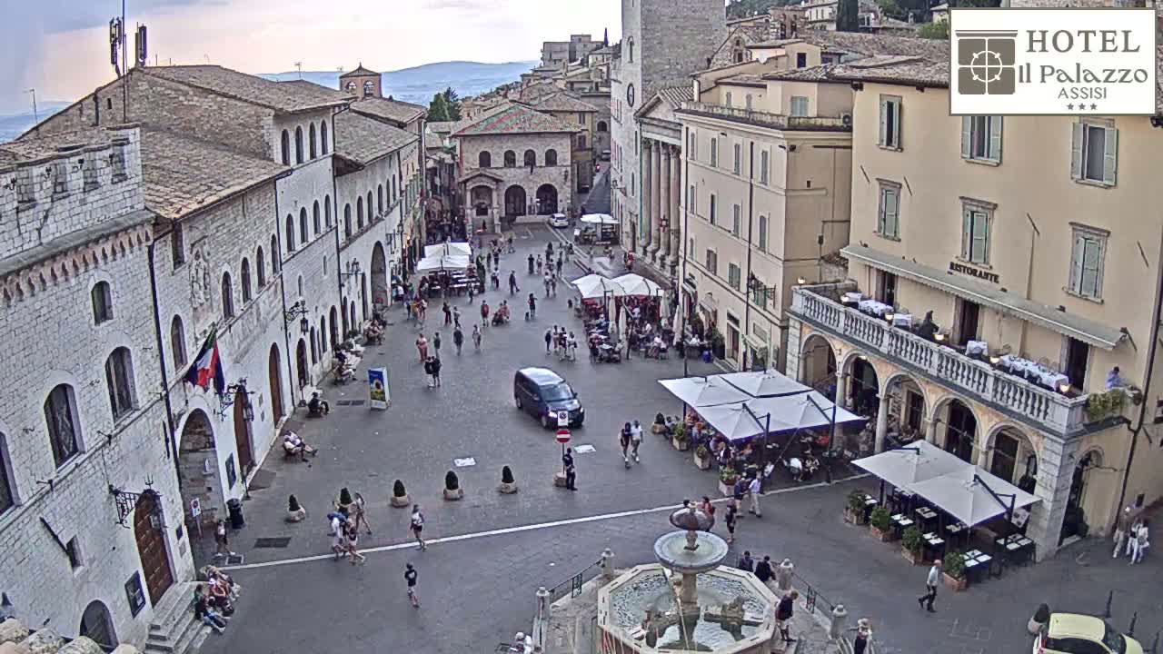 A sunny day reveals a large town square with several buildings, outdoor seating areas under umbrellas, and a fountain in the center.