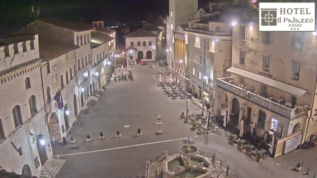 This nighttime aerial view captures a historic town square illuminated by streetlights, surrounded by traditional buildings with outdoor dining areas, under a clear, dark sky.