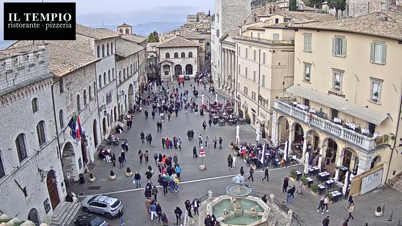 A bustling European town square is viewed from above on an overcast day, filled with numerous pedestrians, outdoor diners, and historic buildings surrounding a central octagonal fountain.